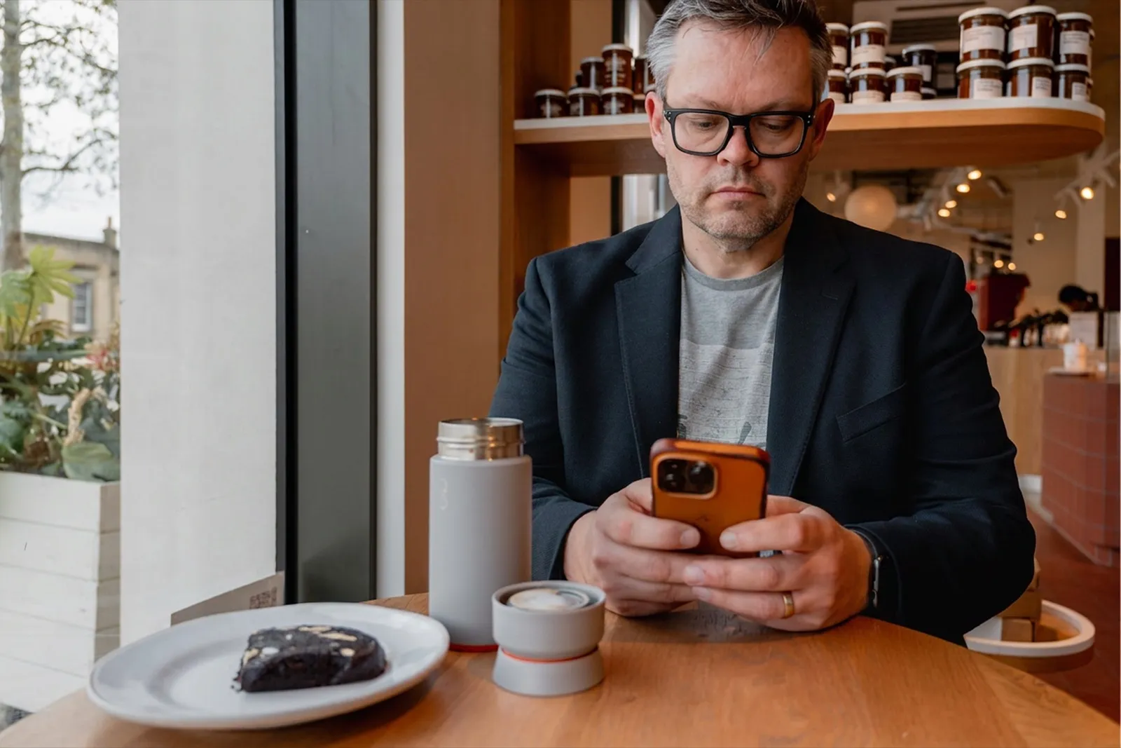 Alistair Stead reading his phone at a cafe table by a window, a flask and a coffee in front of him